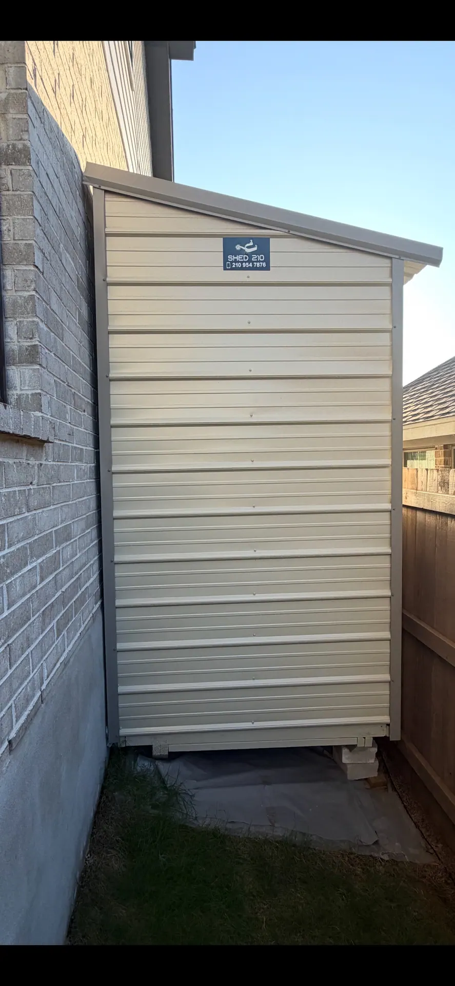 Rear view of the narrow cream metal shed fitted between a brick home and wood fence in Bulverde, TX