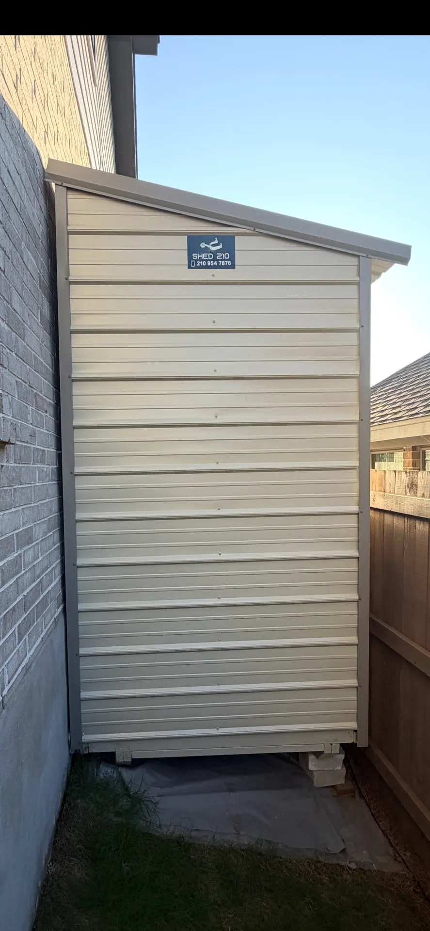 Straight-on rear view of the cream metal shed with tan trim and single-slope roof beside a brick wall