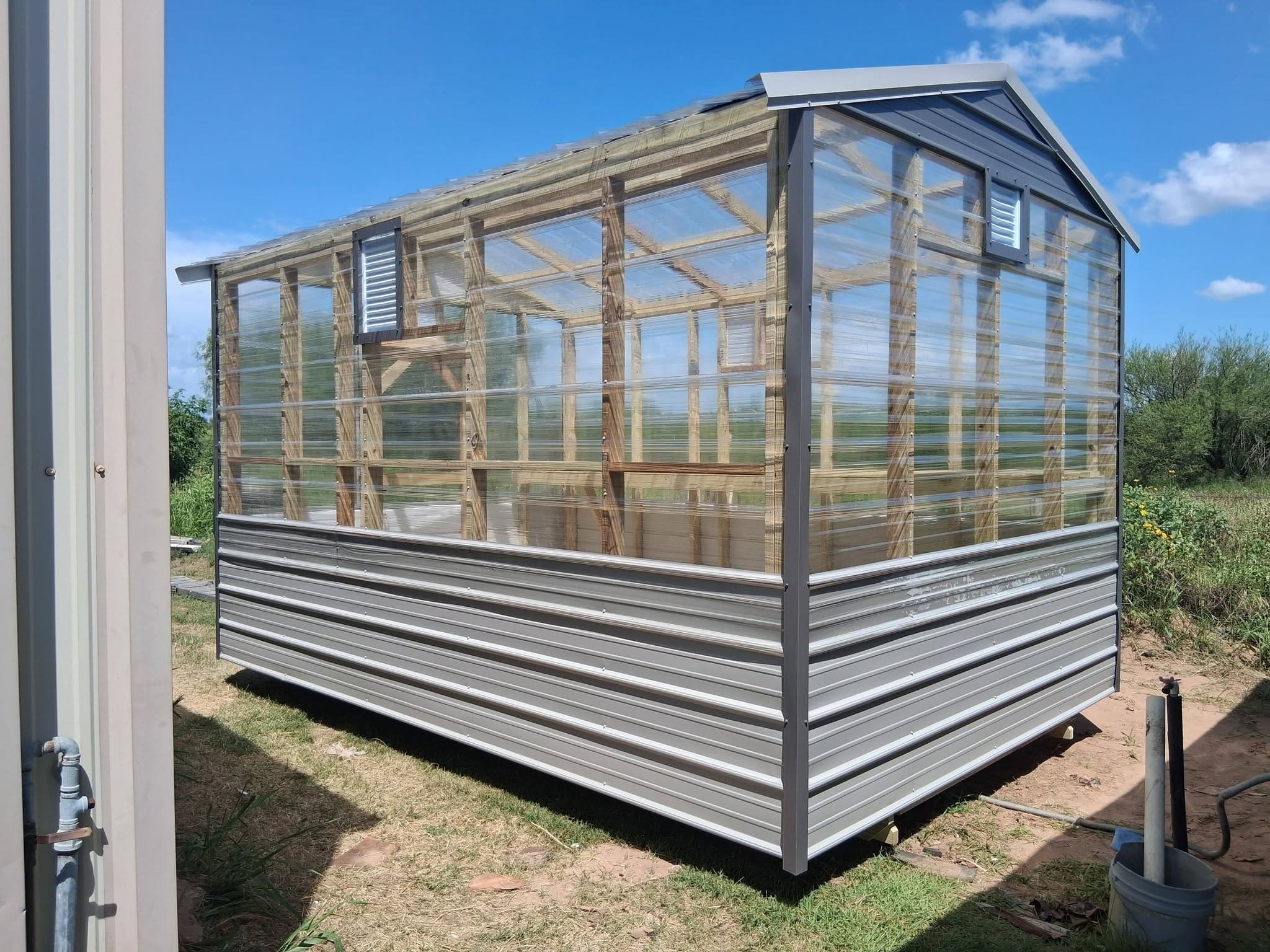 Back and side exterior of the 8x12 greenhouse with clear panels, metal lower walls, and louvered vents