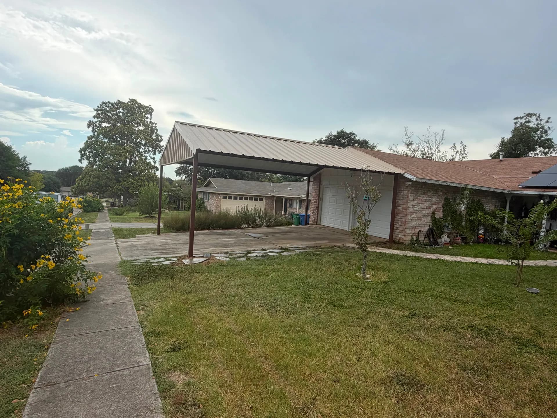 Wide side view of the welded red-iron carport covering both bays of a residential driveway.