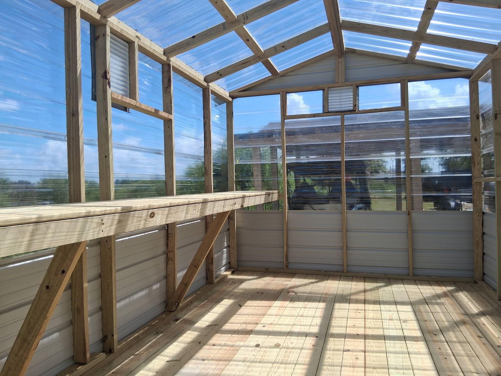 Wide greenhouse interior with a solid wood floor, built-in planting bench, clear panels, and vents
