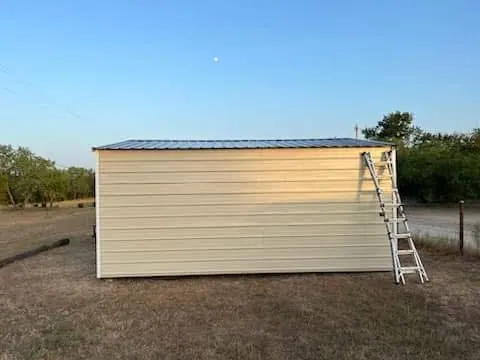 Long side elevation of the completed light tan metal shed with its gable roof and a ladder at the rear corner.