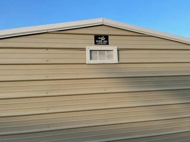 Close view of the shed’s rear gable showing a white-trimmed vent and Shed210 badge beneath the light metal roof.
